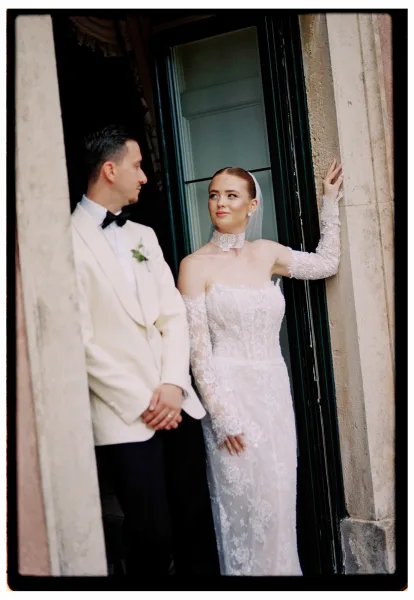 Couple portrait of bride in long-sleeve lace dress and veil gazing at groom in white tuxedo by a stone column doorway entrance