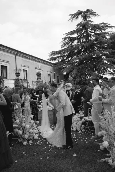 Wedding kiss as bride in a long veil and groom in a suit embrace on a petal-strewn aisle with guests and white florals in a garden villa setting