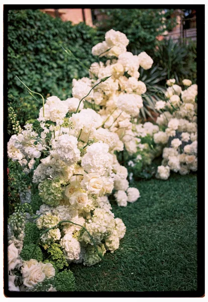 Wedding aisle flowers with white hydrangeas and roses accented by greenery, lining a grass lawn ceremony walkway beside a hedge wall