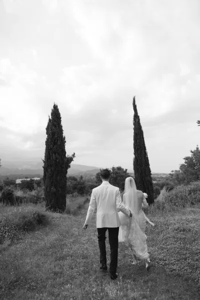 Couple portrait of bride and groom walking away hand in hand, veil trailing, across a grassy field with cypress trees and hills