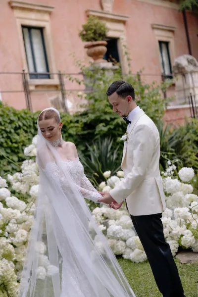 Couple portrait of bride and groom holding hands, her long veil and lace sleeves beside white florals on a garden lawn by pink stucco building