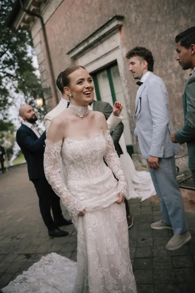 Bride portrait in a strapless lace wedding dress with off-the-shoulder sleeves, laughing in a stone courtyard near groomsmen