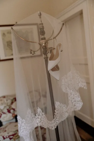 Bridal veil with lace edge hanging on a coat rack beside white pointed-toe heels in a refined room with framed art and molding