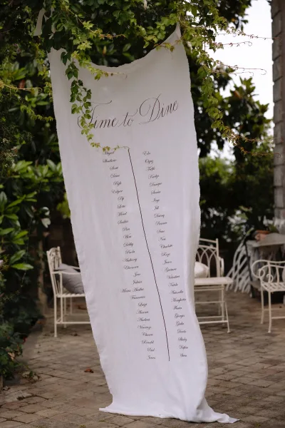 Wedding seating chart on linen fabric with calligraphy lettering, hanging from ribbon ties with greenery vines on a garden patio backdrop