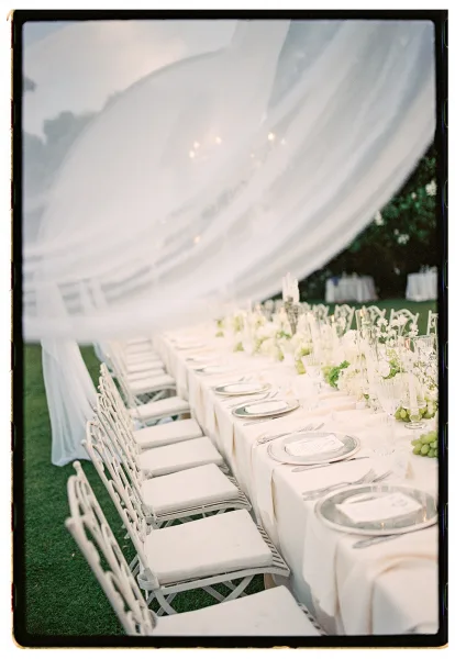 Reception tablescape with an outdoor reception table set in white linens, crystal glassware, taper candles, and greenery garland under string lights