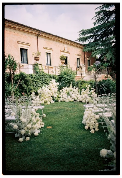 Ceremony aisle design with white floral ground arrangements lining a lawn aisle, metal chairs set before a villa and garden greenery
