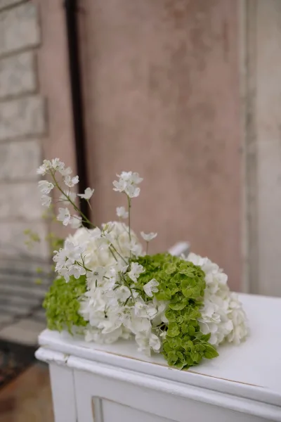 Wedding floral arrangement with green hydrangea and white flowers on a white dresser, set against a brick wall and wooden door backdrop