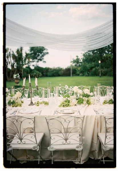 Reception tablescape with an outdoor wedding reception table under white drapery canopy, silver candelabra, white florals, and grapes on a lawn