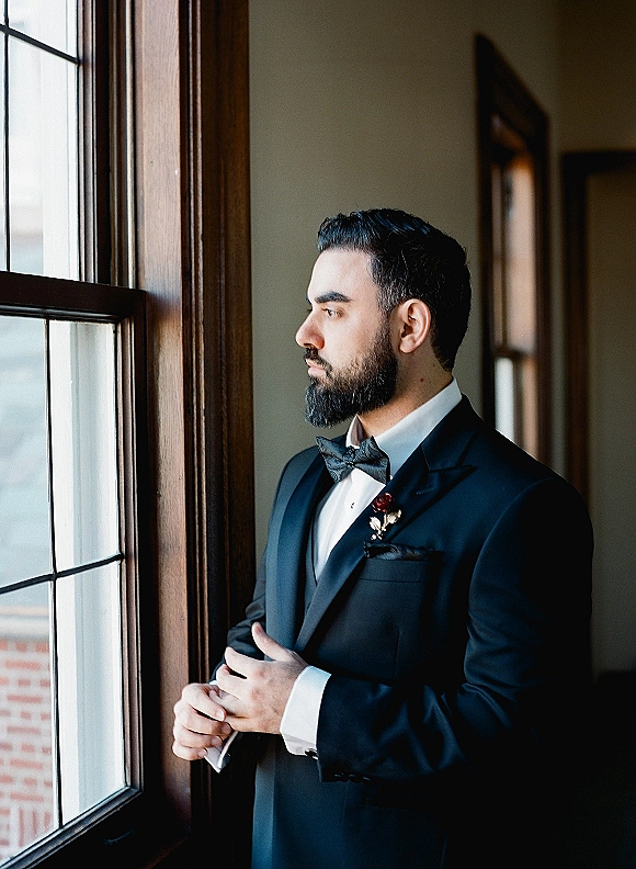 Groom portrait in a black tuxedo with bow tie, boutonniere, and pocket square, standing by a window in soft natural light indoors