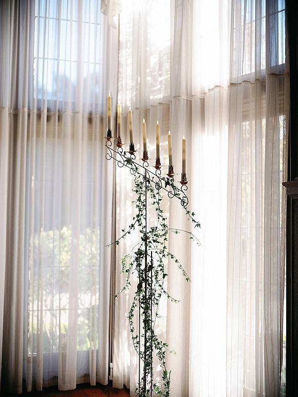 Wedding candelabra taper candle centerpiece with white tapers and trailing ivy on a metal stand by sunlit tall windows and sheer curtains