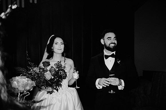 Wedding reception toast as bride and groom raise champagne flutes, bride holding bouquet, guests silhouetted against dark drapery backdrop