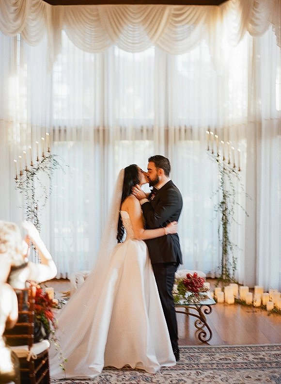 Wedding kiss as bride in a long veil and strapless gown meets groom in a black suit beneath a candlelit arch with sheer drapes and windows