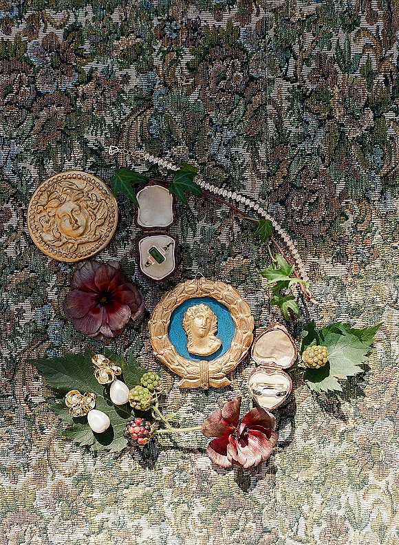 Wedding ring flatlay with wedding rings in ring box, pearl earrings and cameo jewelry on floral tapestry fabric with ivy and berries