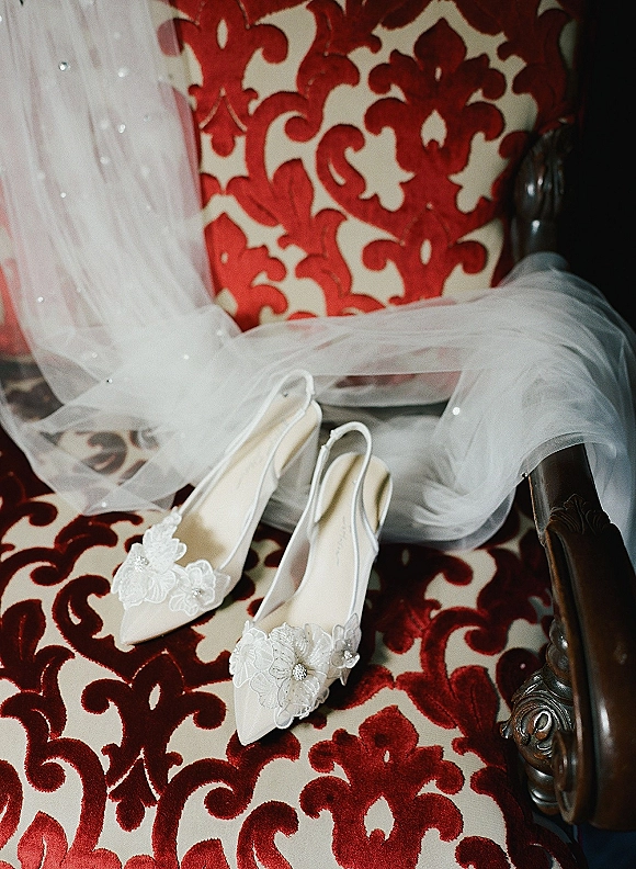 Bridal shoes—white lace wedding heels with floral appliqué and rhinestone sparkle, draped with a tulle veil on a red damask chair
