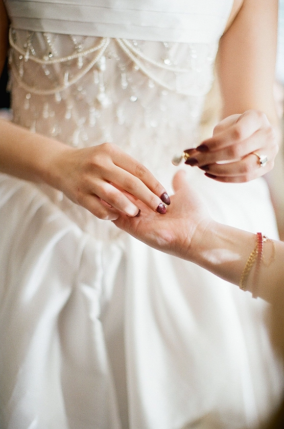 Wedding ring exchange close up as the groom slides a band onto the bride’s finger, showing burgundy manicure, bracelet, and beaded bodice in soft indoor light