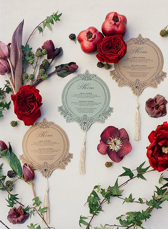 Wedding menu cards in an ornate wedding menu flatlay with tassels, red roses, pomegranates, ivy, and hellebore on light fabric backdrop