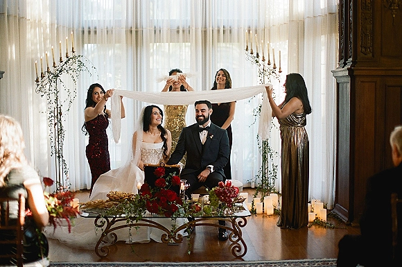 Ceremony moment at an indoor wedding ceremony with bride and groom seated under a chuppah, candlelit with greenery garland and sheer drapery backdrop