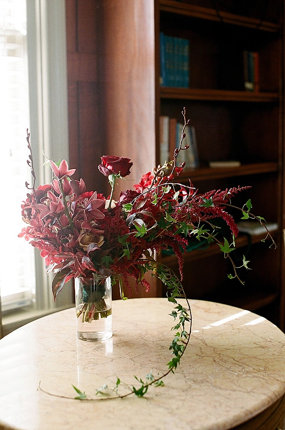 Wedding bouquet of red roses and pink orchids with trailing ivy in a water-filled glass vase on a marble table by bookshelves