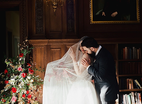 Couple portrait of bride and groom embrace as he kisses her forehead, bridal veil flowing beside red roses in a wood-paneled library nook