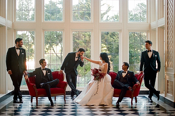 Wedding party portrait with bride in strapless dress seated on red velvet chairs as groomsmen in black tuxedos pose by tall grid windows