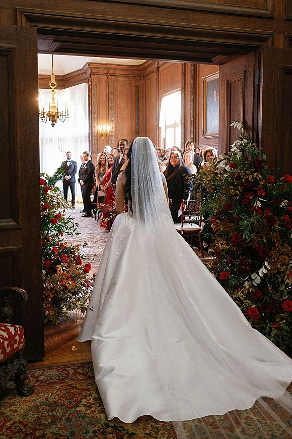 Bridal processional as the bride walking down aisle from behind in a satin ball gown with cathedral veil and red rose florals under a chandelier