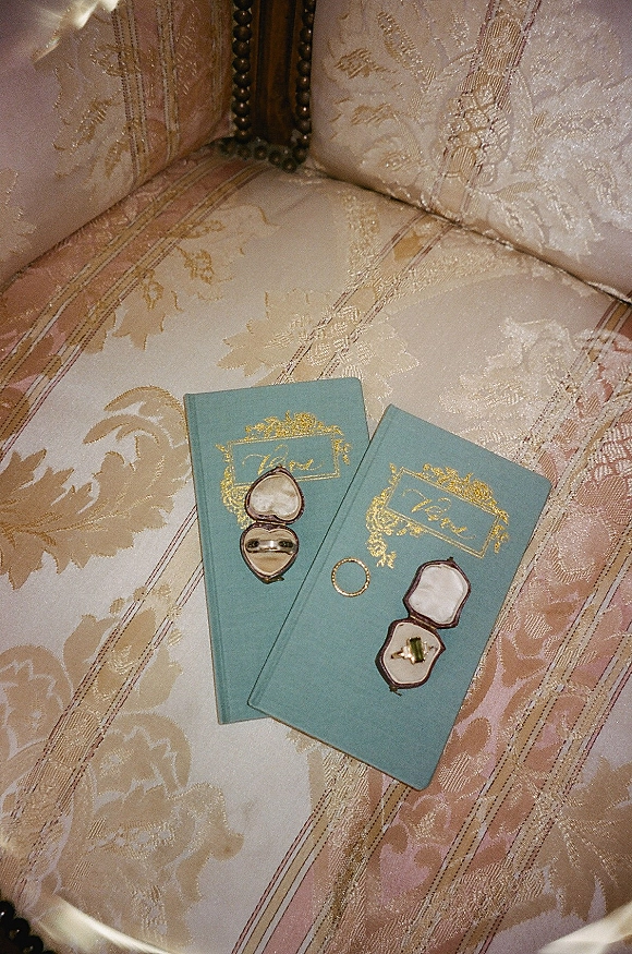 Wedding rings with a gold band and solitaire engagement ring in a velvet ring box beside vow books on a vintage brocade chair