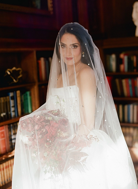 Bridal portrait of a bride with veil over her face holding a deep red bouquet in a wood-paneled library lined with bookshelves