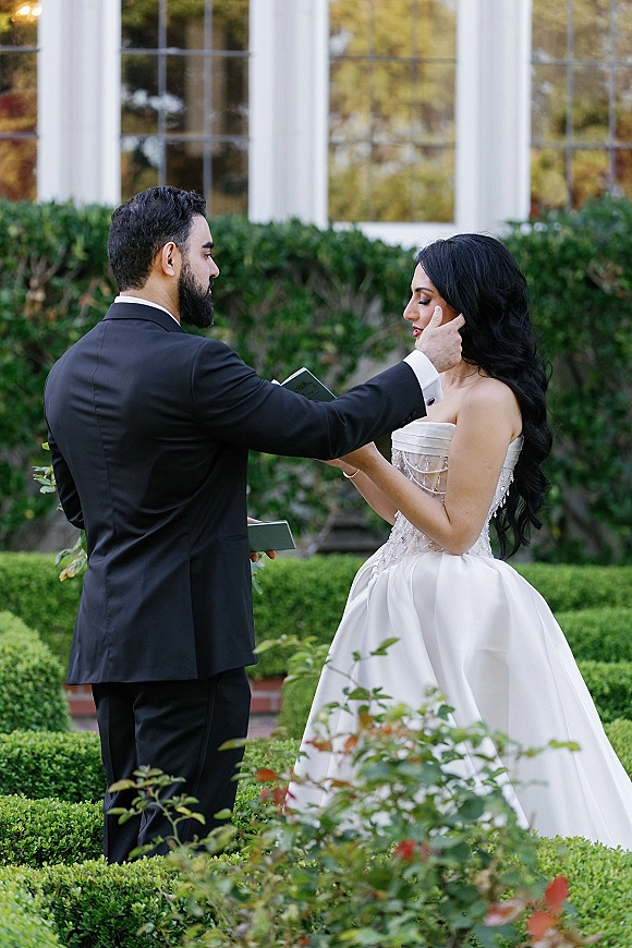 Wedding vows as groom reads from vow booklets and touches bride’s face, her strapless beaded gown framed by garden hedges and brick steps