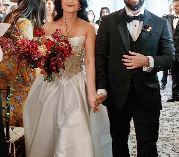 Recessional moment as bride and groom walk down the aisle holding hands, her red bouquet and strapless dress amid cheering guests indoors