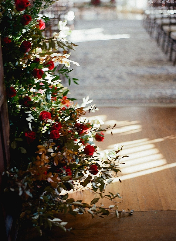 Ceremony aisle flowers with red rose aisle arrangement and lush greenery on a wood floor, leading to chair rows in sunlit shadows