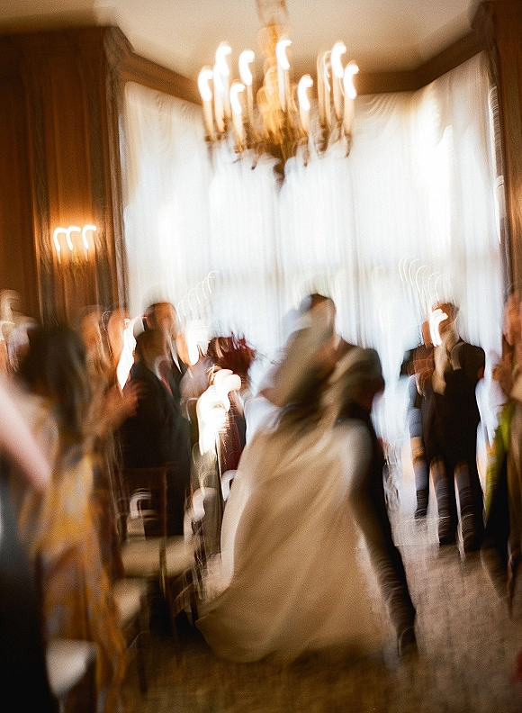 First dance as groom spins bride in flowing wedding dress, bouquet in hand, under chandelier in wood-paneled ballroom with window light