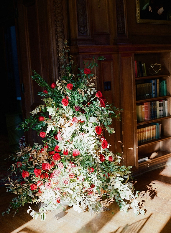 Wedding floral installation with red roses, white blooms, and greenery garland spread on the floor against wood-paneled library walls with bookshelves