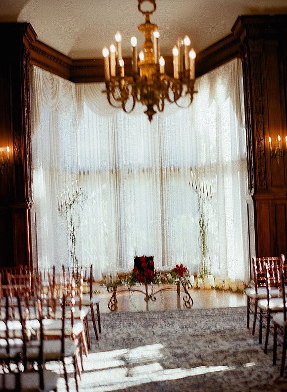 Ceremony setup for an indoor wedding ceremony with wood chairs lining an aisle runner, white drapery, candles, and chandelier by tall windows