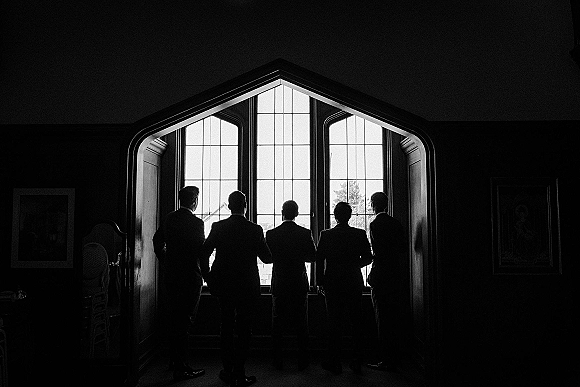 Groomsmen portrait with suited men in dress shoes standing by an arched window in an interior room with wall art and chairs