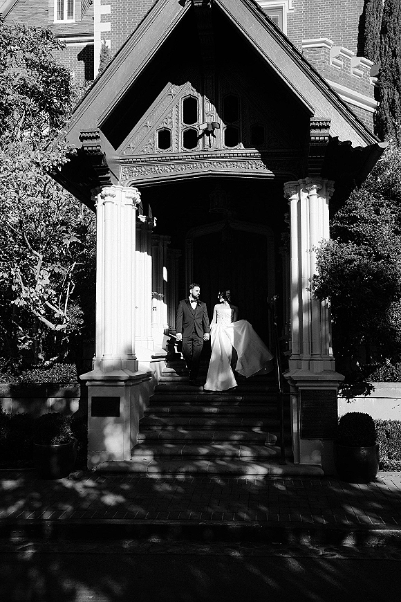 Couple portrait in a black and white wedding portrait style, bride and groom holding hands on stone steps by a columned entryway