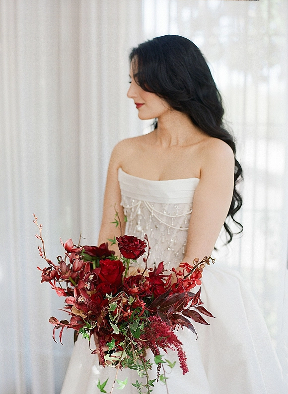 Bridal portrait of a bride holding bouquet, wearing a strapless beaded gown and red lipstick by sheer curtains in window light