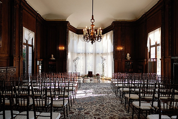Indoor ceremony setup in a library wedding ceremony with chiavari chairs, taper candles, floral aisle decor, and chandelier lighting in a wood-paneled room