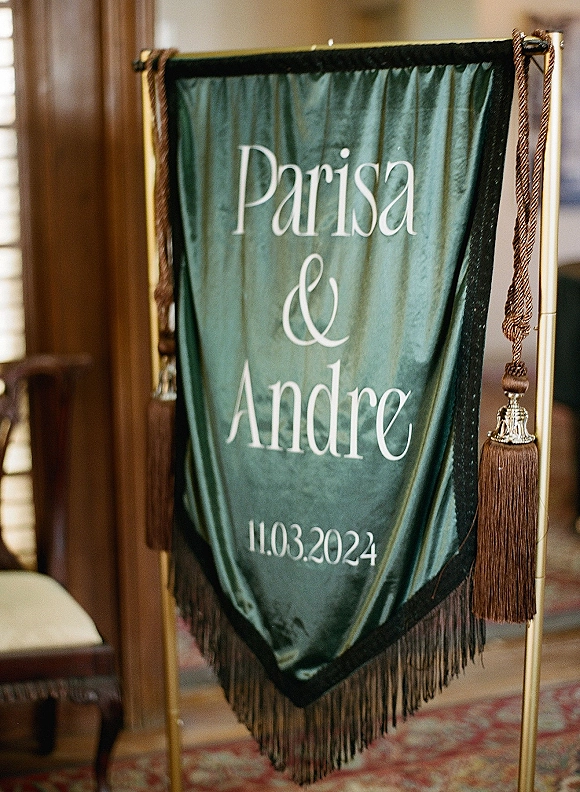 Wedding welcome sign with personalized wedding sign details on a fabric banner with fringe trim, hanging on a gold stand in a wood-paneled room