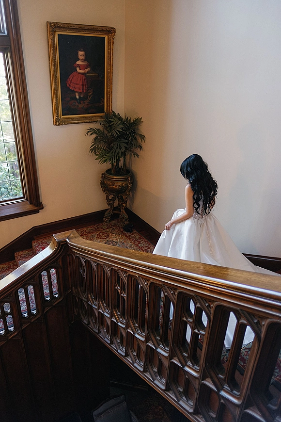 Bridal portrait of a bride on staircase in a strapless corset-back gown, long dark hair flowing, window light in a cream-walled stairwell