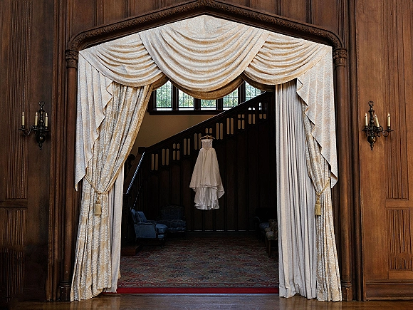Wedding dress hanging in an arched doorway, bridal gown on hanger framed by draped curtains and wood-paneled hallway with sconces