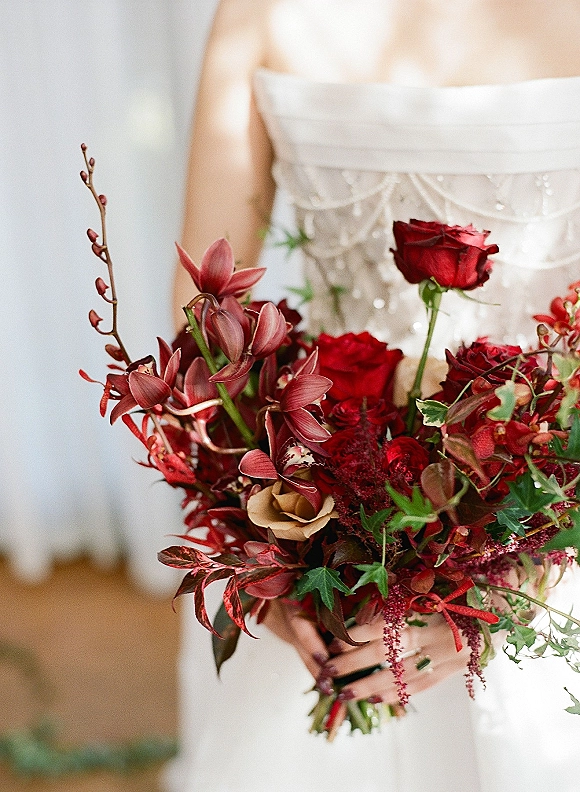 Bridal bouquet with red rose blooms and burgundy flowers, orchids, greenery, and ribbon held against a strapless dress indoors