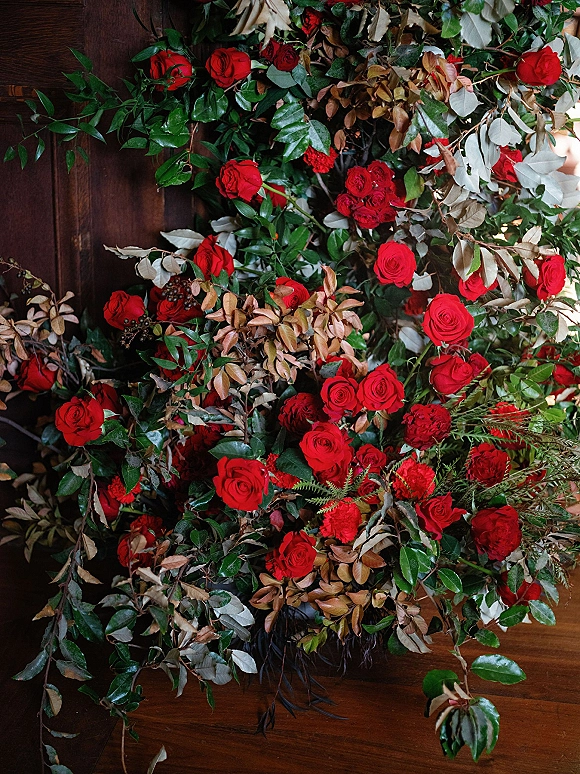Wedding floral installation of red rose floral arrangement with lush greenery and ferns cascading over a rustic wooden wall backdrop