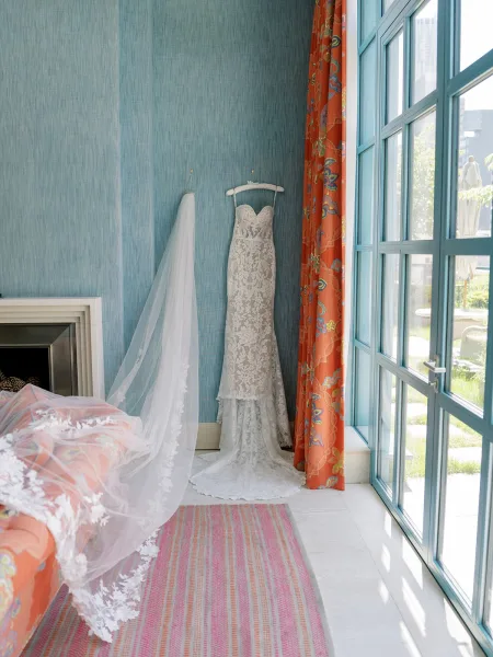 Wedding dress with lace wedding dress detail hanging by a bright window, long veil draped over the train in a blue-walled suite