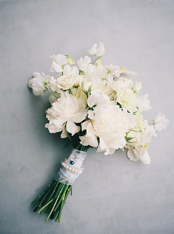 White bridal bouquet of white peonies with ribbon wrap and a blue gemstone charm, styled as a flat lay on a gray surface