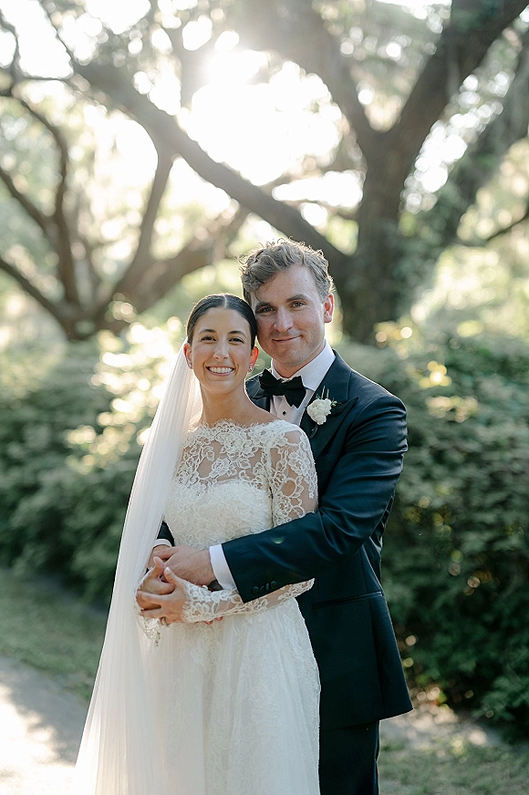 Couple portrait of bride and groom smiling in a newlywed embrace, her lace-sleeve dress and veil glowing under sunlit trees