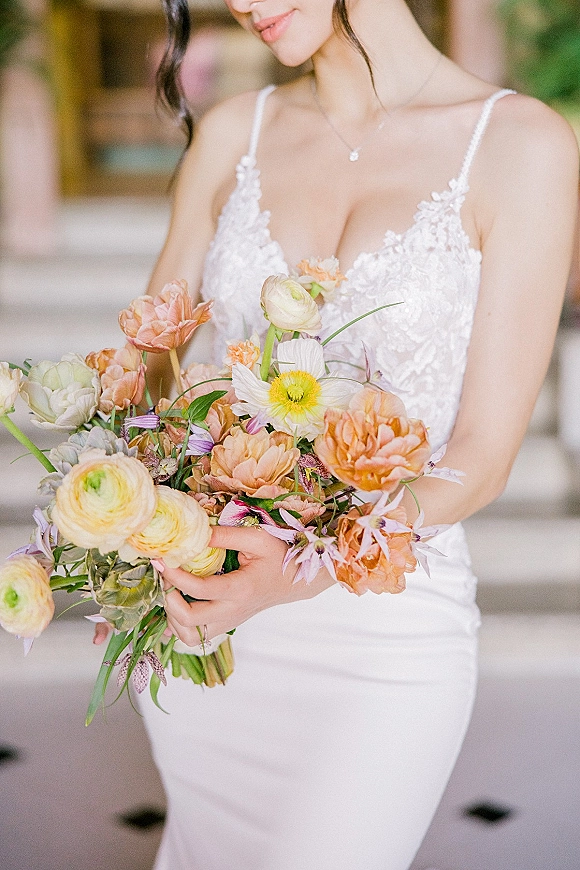 Bridal portrait of a bride holding bouquet of pastel ranunculus with a lace spaghetti-strap gown and pendant in a soft indoor setting