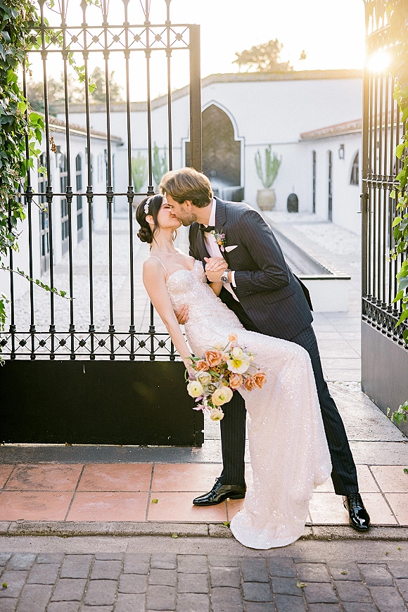 Wedding kiss portrait of groom dipping bride in a dip kiss wedding pose by an iron gate, bride in strapless sequin dress holding bouquet in sunset light