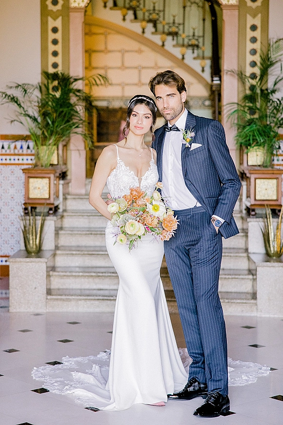 Couple portrait of bride holding bouquet beside groom in navy pinstripe suit on a grand staircase with potted palms indoors