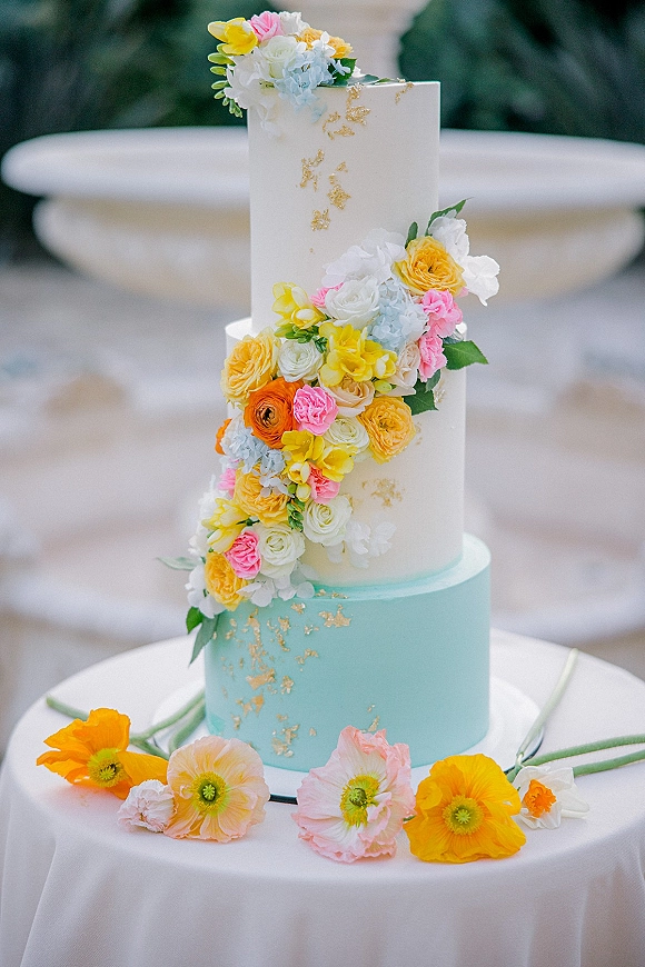 Wedding cake with three tier wedding cake design, sugar flower floral cascade and gold leaf on a stand outdoors by blurred greenery