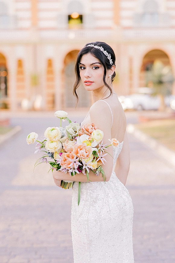 Bridal portrait of an over-the-shoulder bride holding bouquet in a beaded strapless gown, standing by an arched courtyard facade